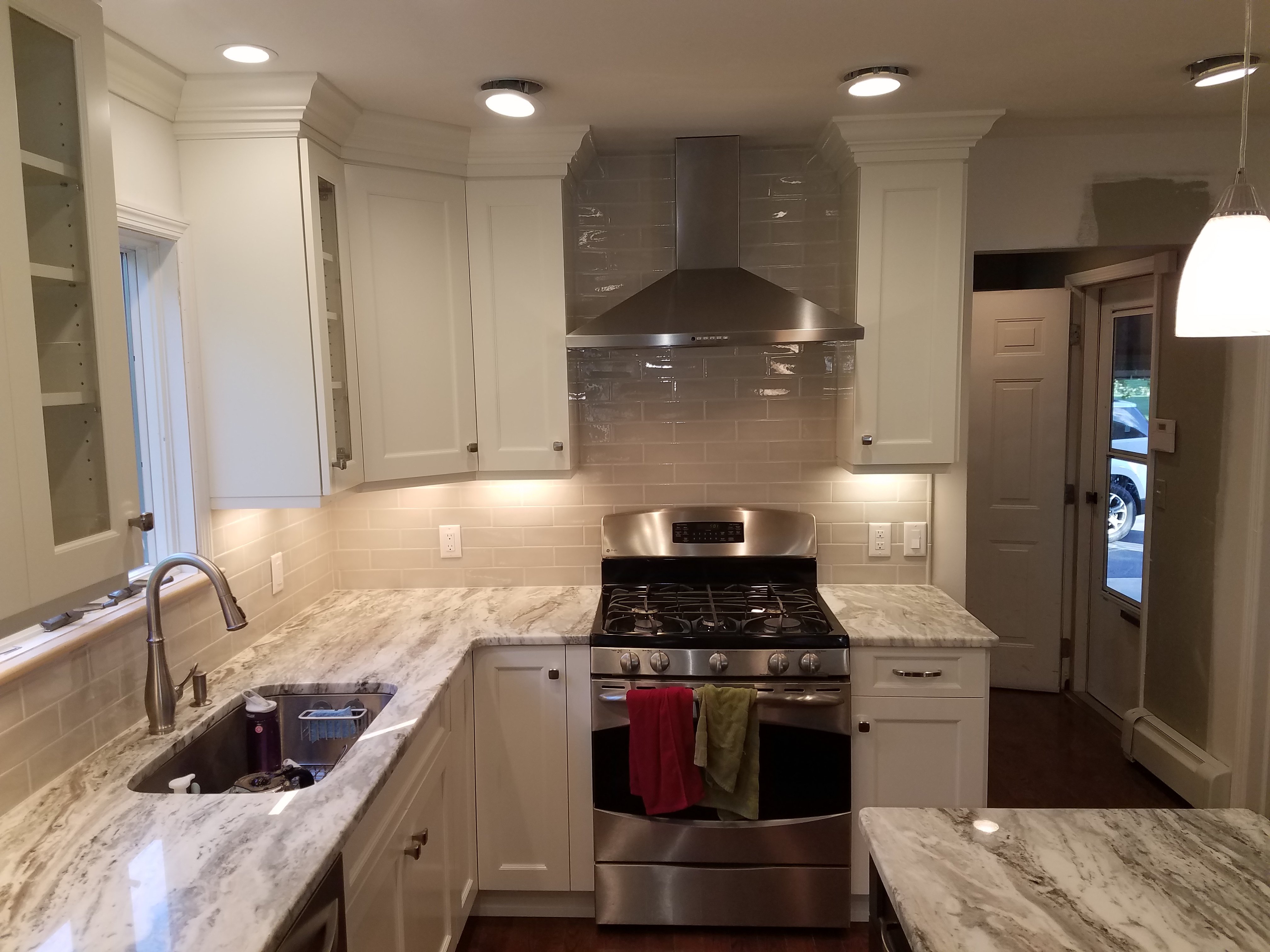 A six-burner stainless steel gas range and matching hood stand against a light gray subway tile backsplash, framed by white shaker cabinets and gray-veined quartz countertops.
