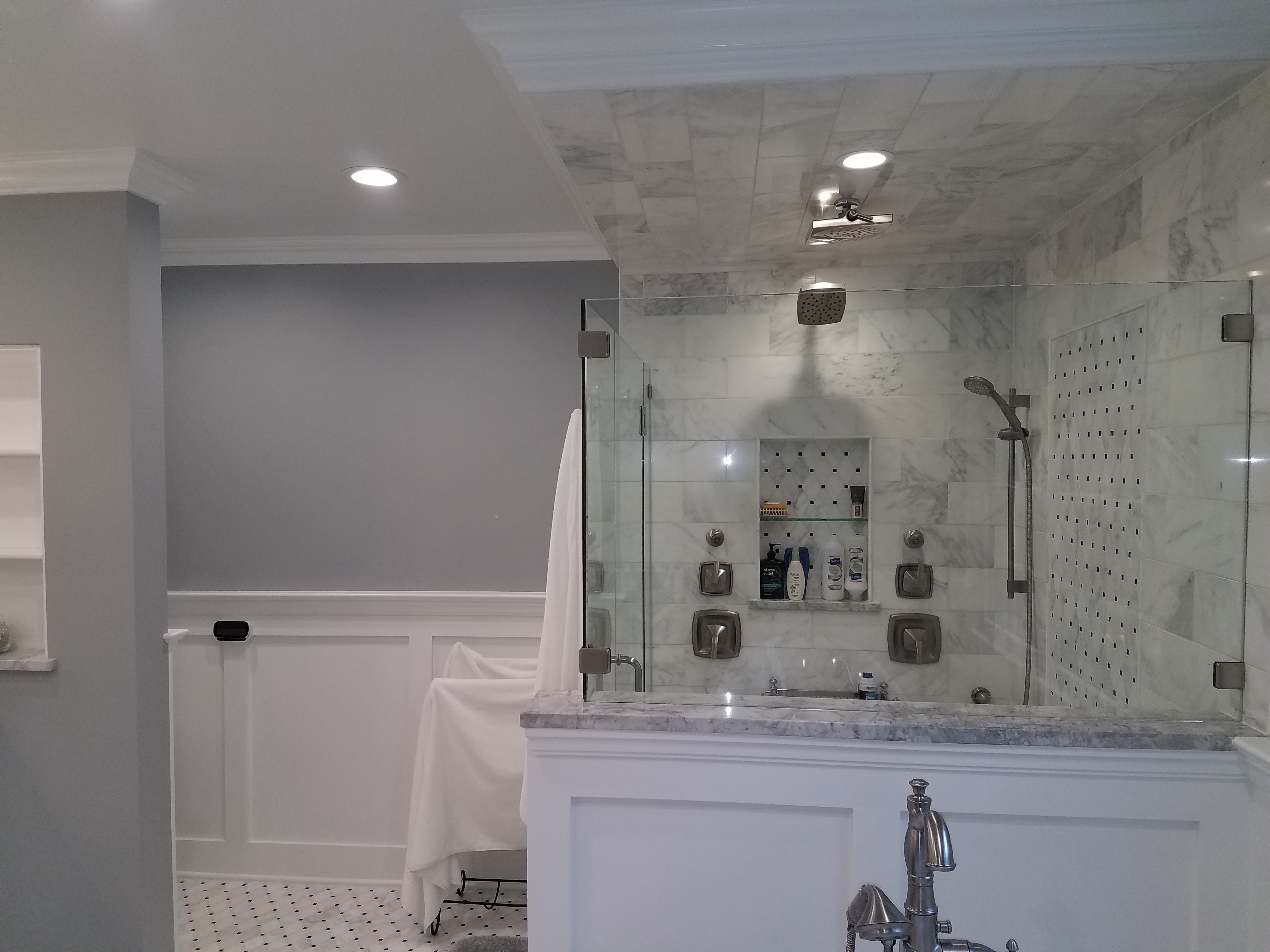 A frameless glass shower has marble subway tile walls, a ceiling-mounted rain shower head, and a tiled storage niche, with white wainscoting in the foreground.