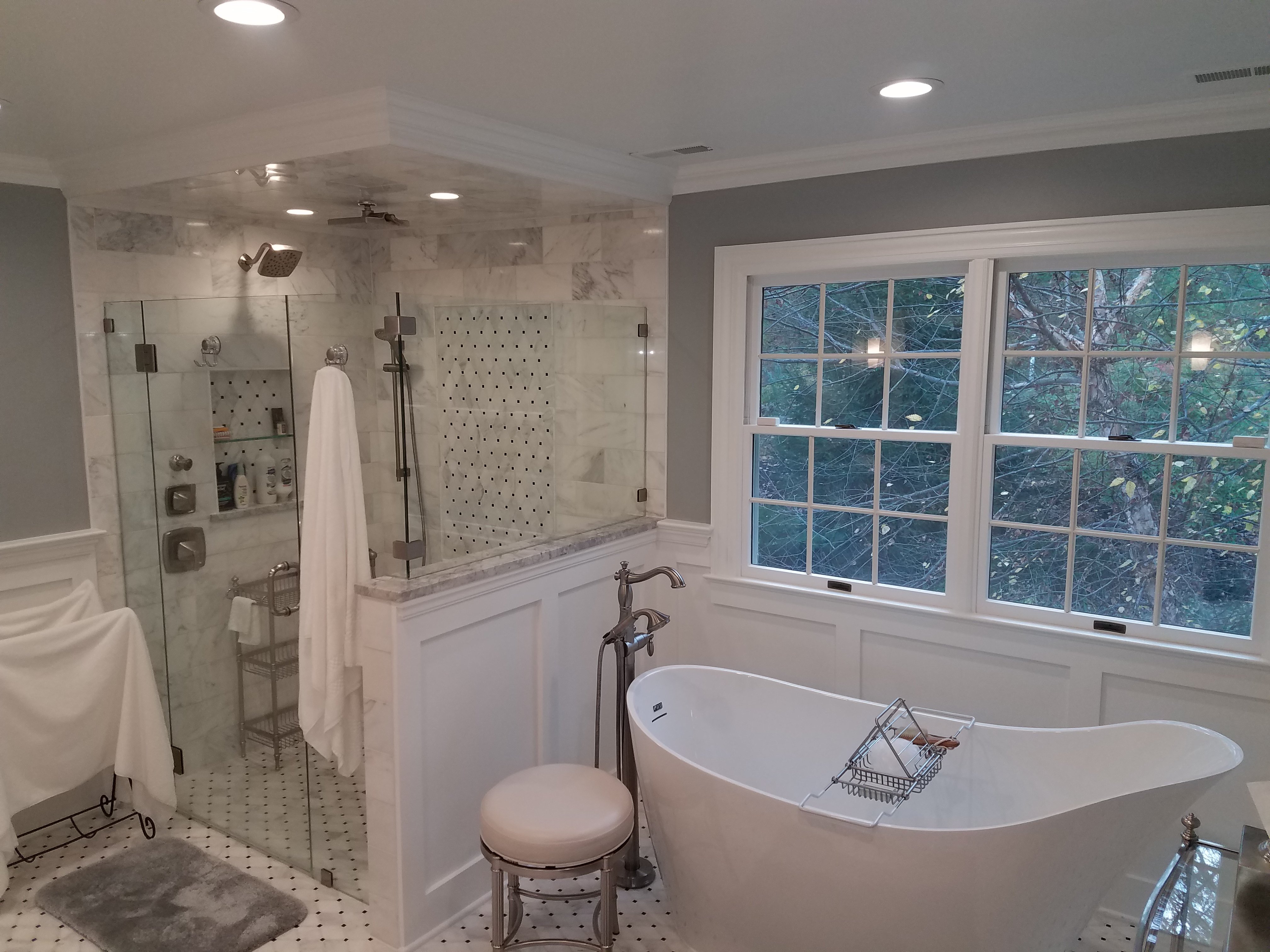 A freestanding white soaking tub sits beneath a window overlooking trees, next to a glass-enclosed marble tile shower with dual shower heads.