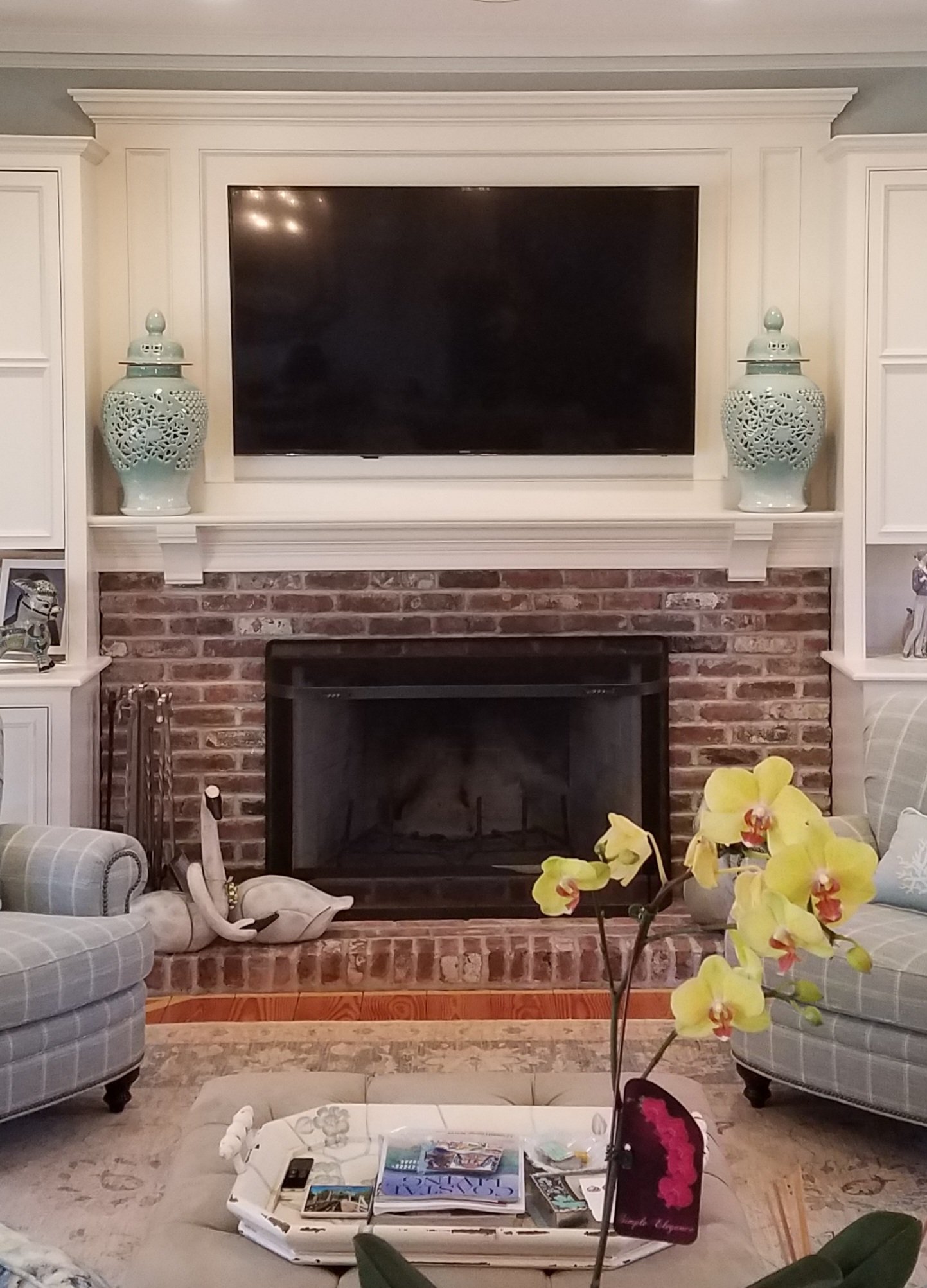 A red brick fireplace with a white wood mantel and mounted flat-screen TV is flanked by gray plaid armchairs over hardwood flooring.
