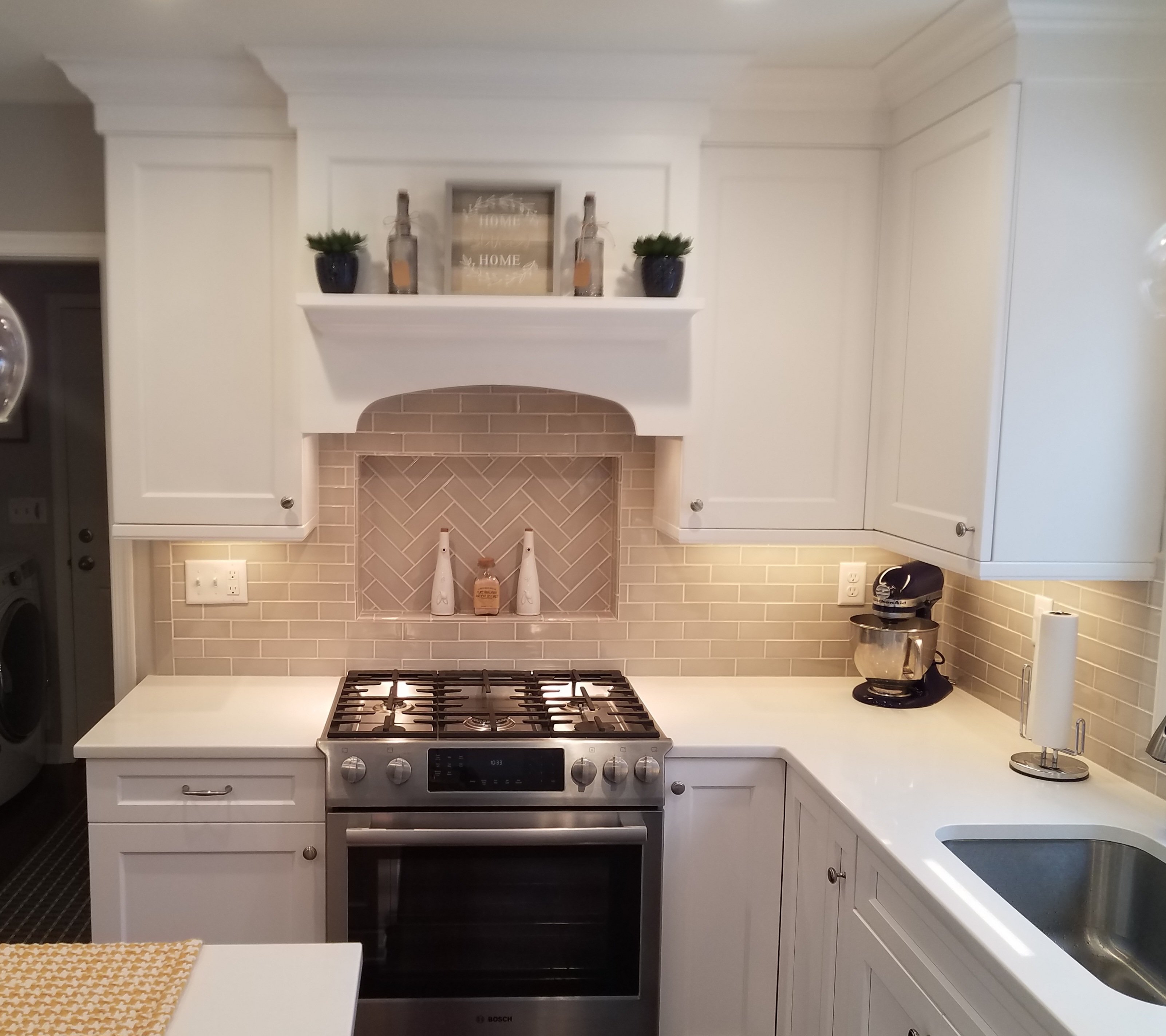 White shaker cabinets surround a stainless steel range and oven, with a tan subway tile backsplash and a herringbone-tile niche above the cooktop.