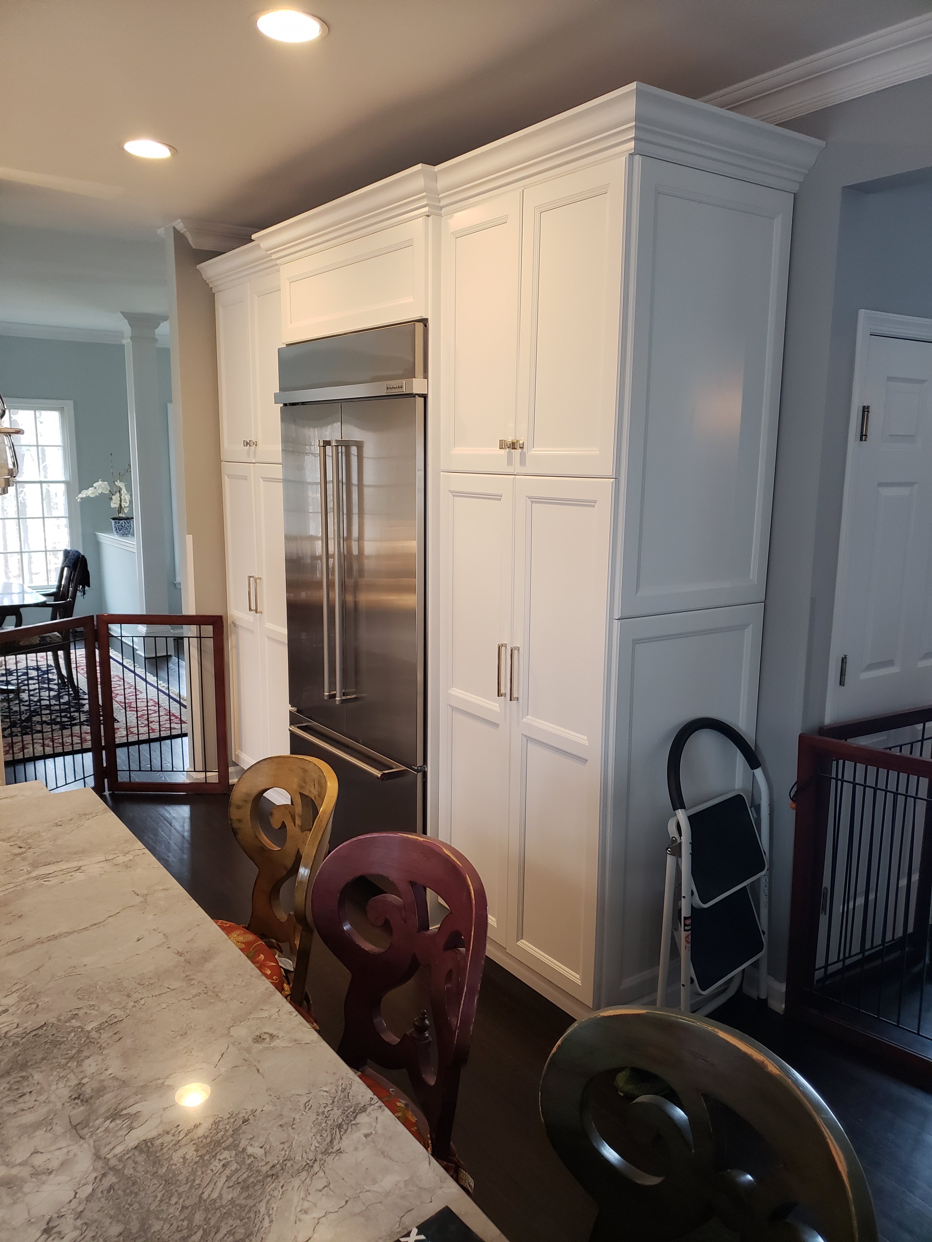 A stainless steel french door refrigerator is built into a wall of white shaker cabinets, with a marble countertop edge and a carved wood chair in the foreground.
