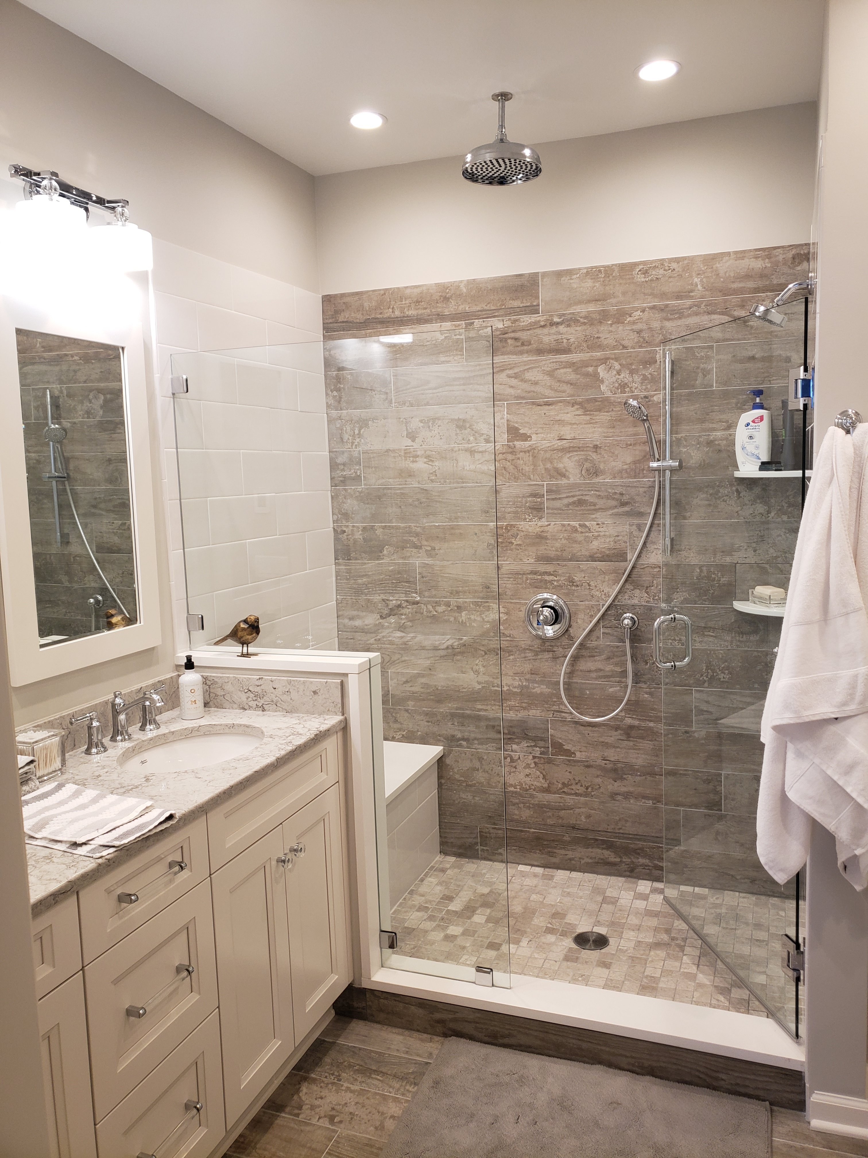 A glass-enclosed walk-in shower has wood-look plank tile walls, a chrome rain shower head, a handheld sprayer, and a built-in tile bench seat.