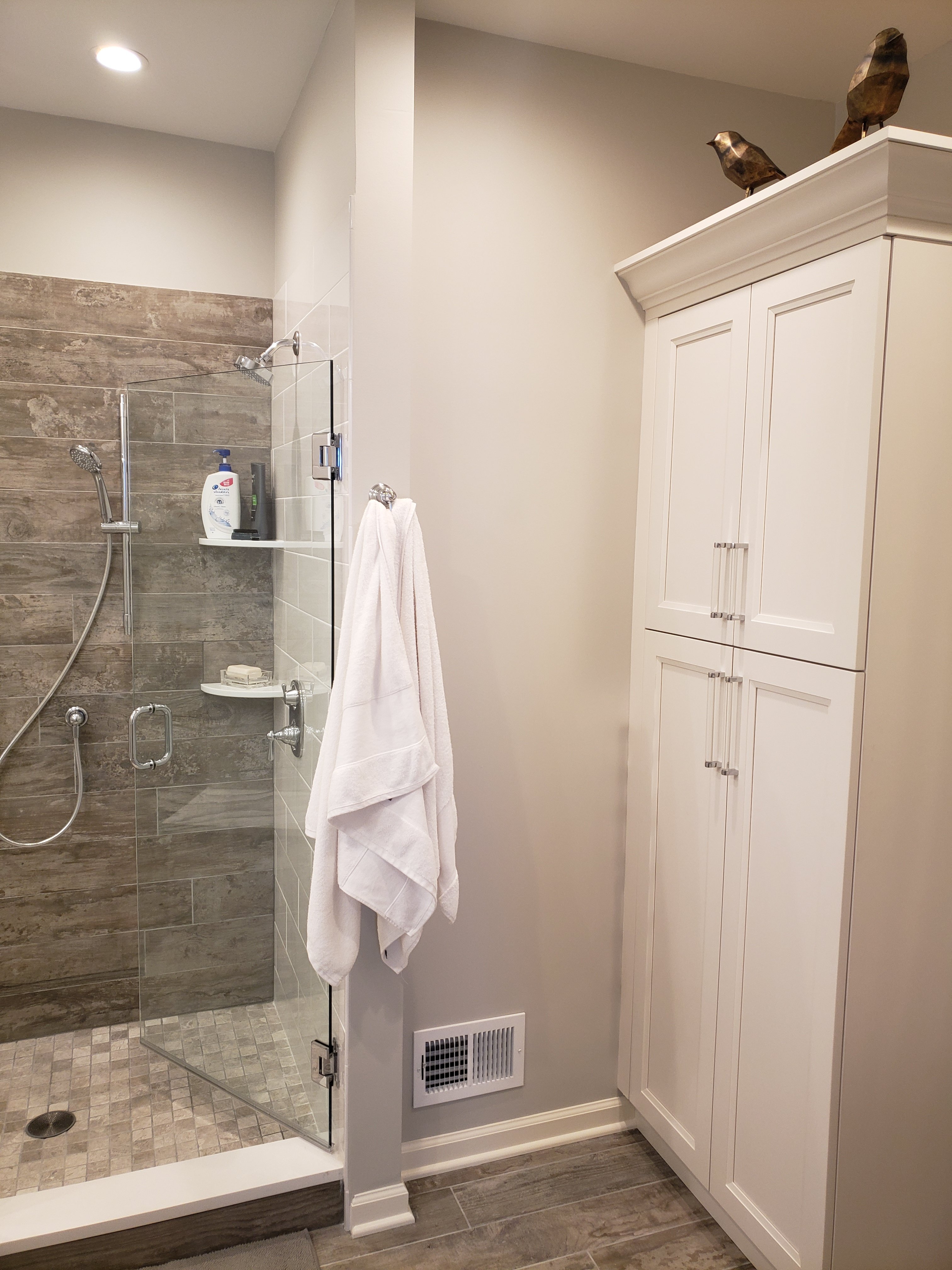 White shaker cabinets with brushed nickel pulls hang above a glass shower door with folded towels on a bar below, and wood-look plank tile flooring inside.