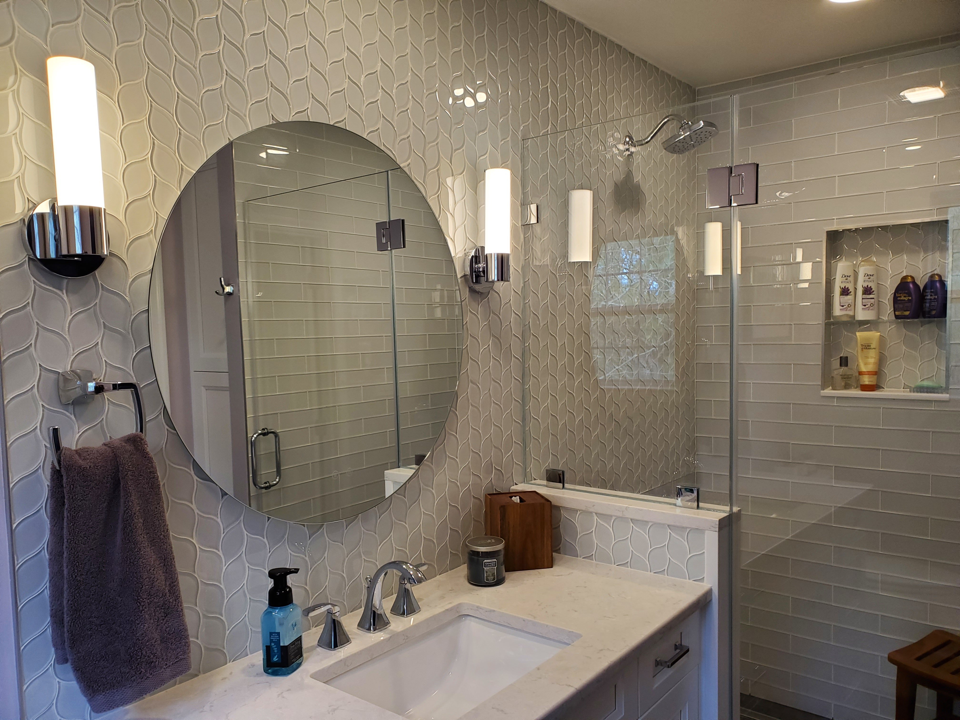 A round frameless mirror hangs above a white quartz vanity with an undermount sink and chrome faucet, set against a wall of light gray wavy mosaic tile.