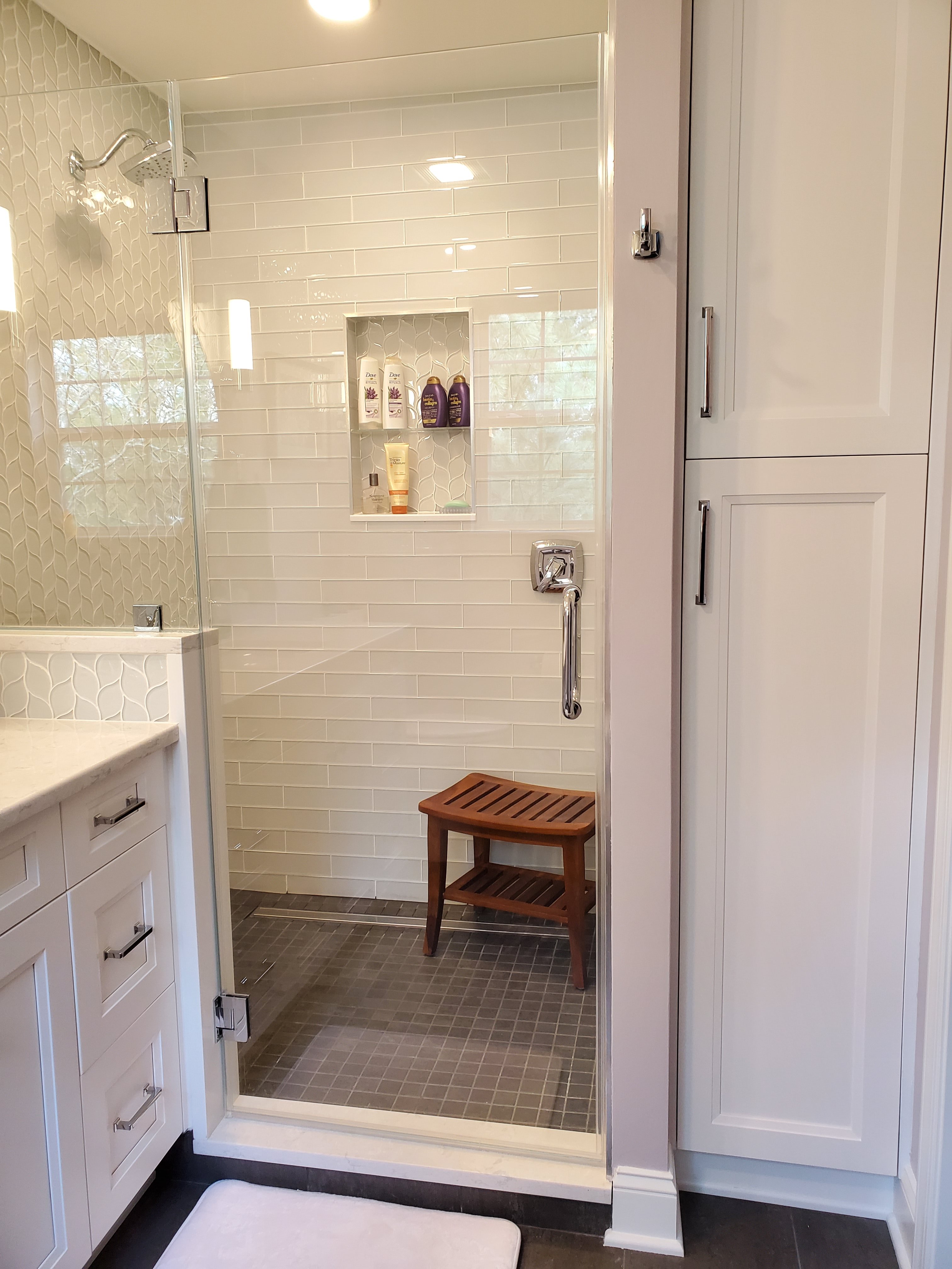 A walk-in shower has light sage green vertical subway tile, a frameless glass door, and chrome fixtures including a grab bar and handheld sprayer.