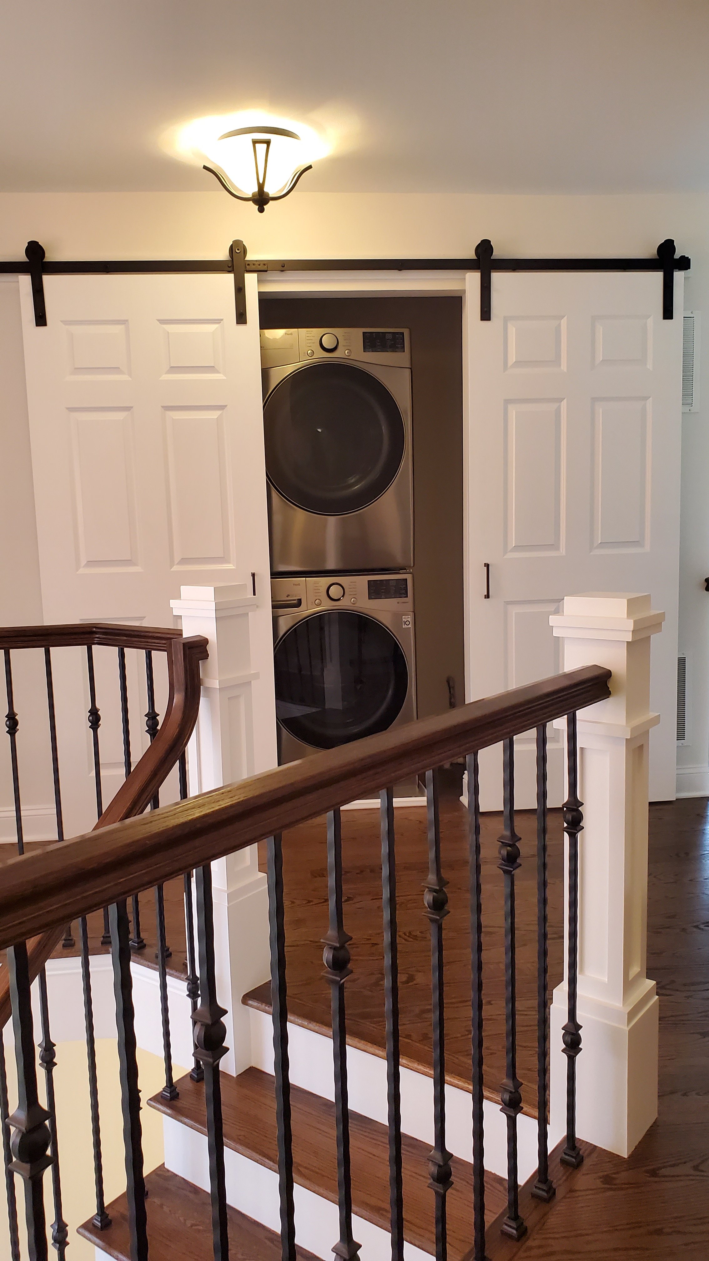 A stainless steel stacked washer and dryer are built into a closet with white paneled doors, positioned beside a staircase with a wood handrail.