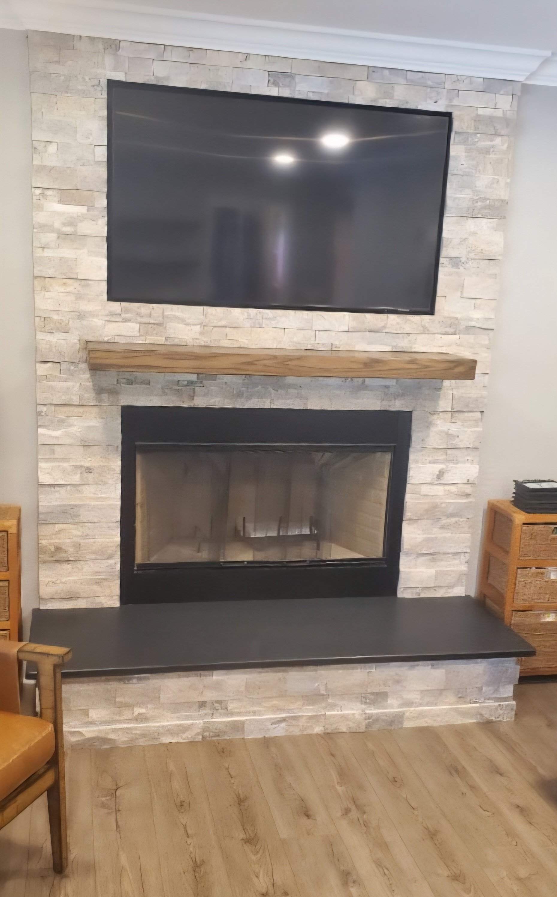 A white and gray stacked-stone fireplace surround has a black metal insert, a wood mantel shelf, and a black stone hearth extending into a bench.