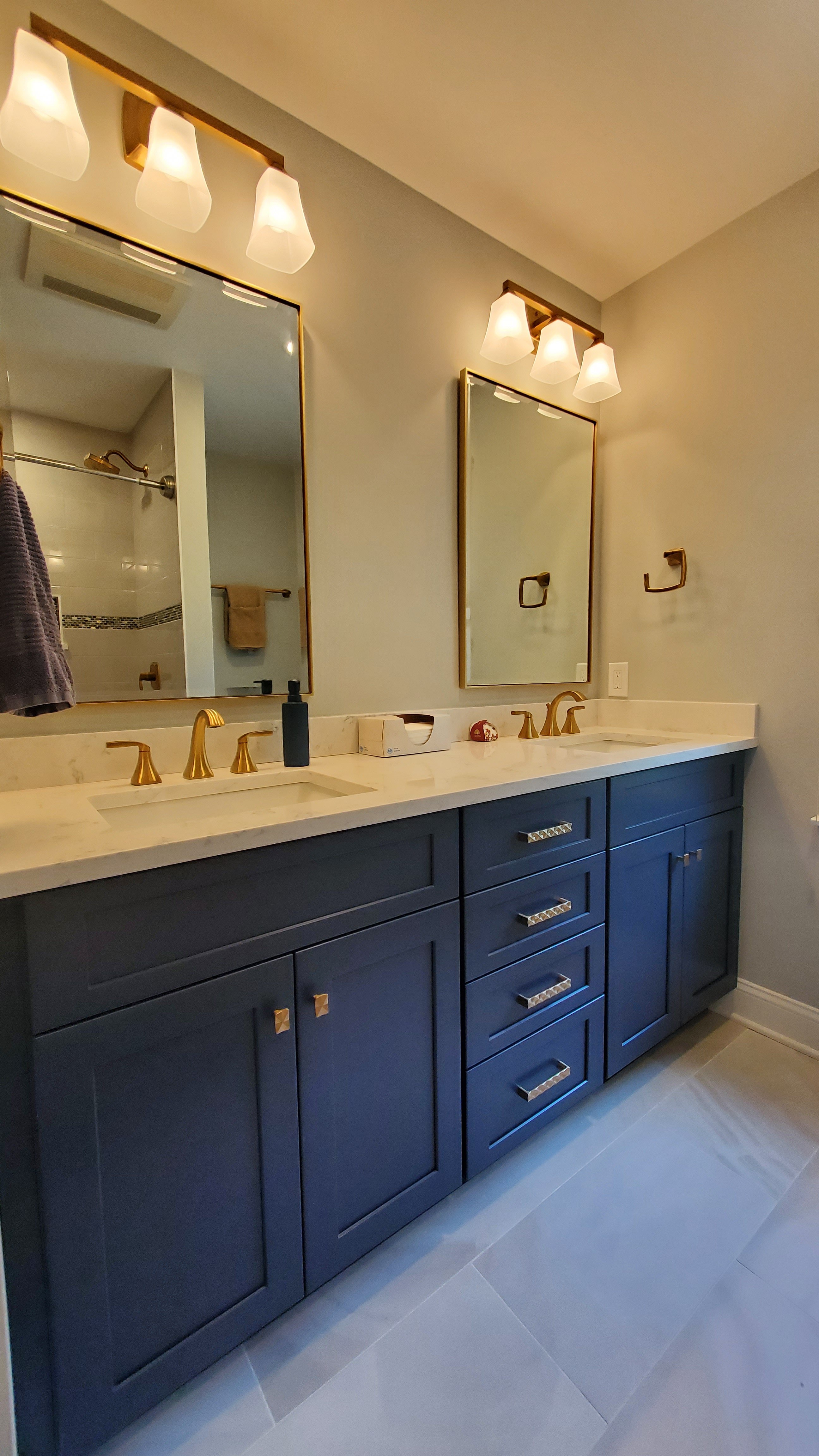 Navy blue shaker-style cabinets with brass hardware line a bathroom wall beneath gold-framed mirrors and glass wall sconces, above a light countertop with brass faucets.
