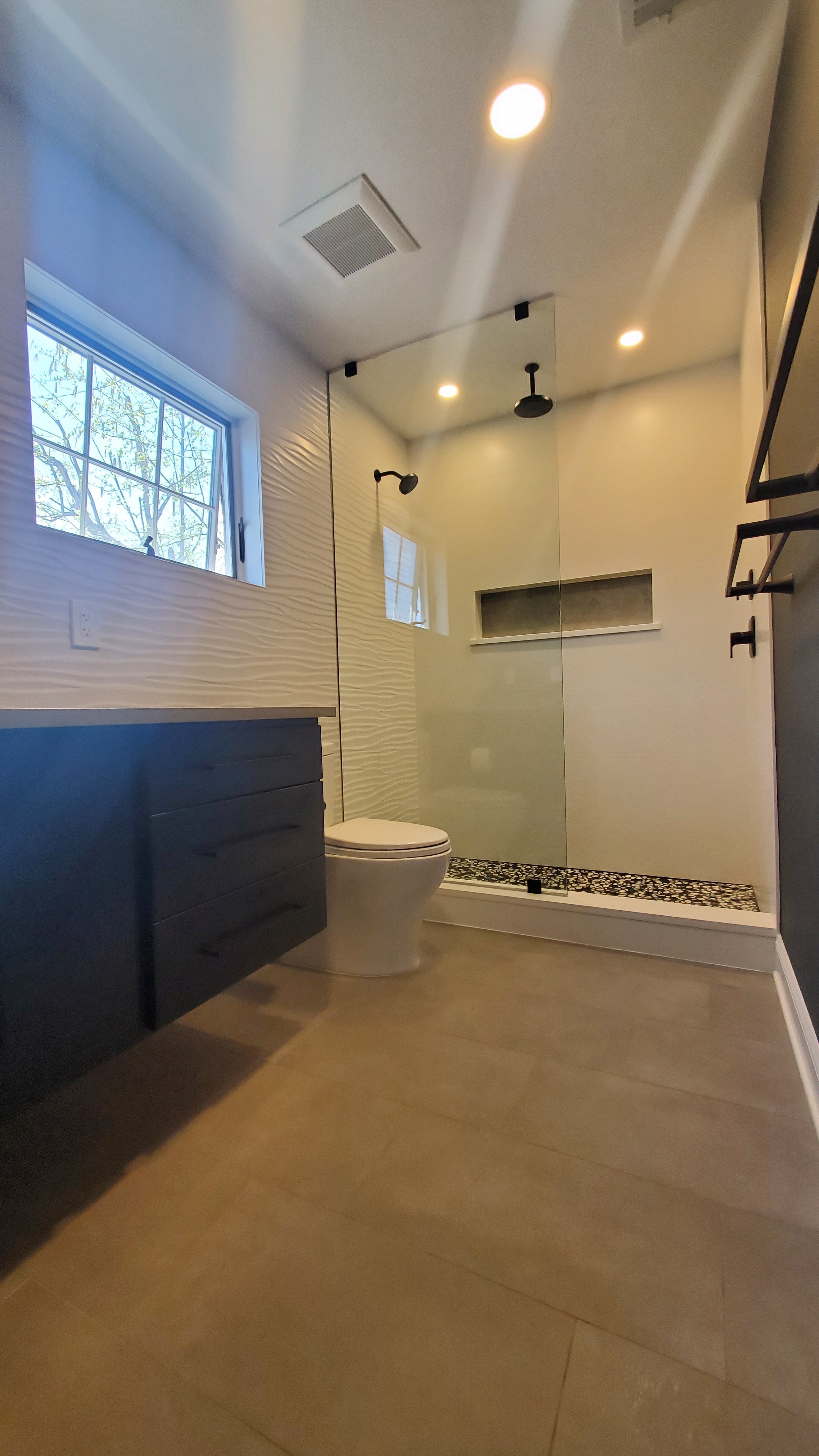 A glass-enclosed shower has large-format tan tile walls, a dark mosaic accent strip, and matte black hardware, beside a white toilet and window.