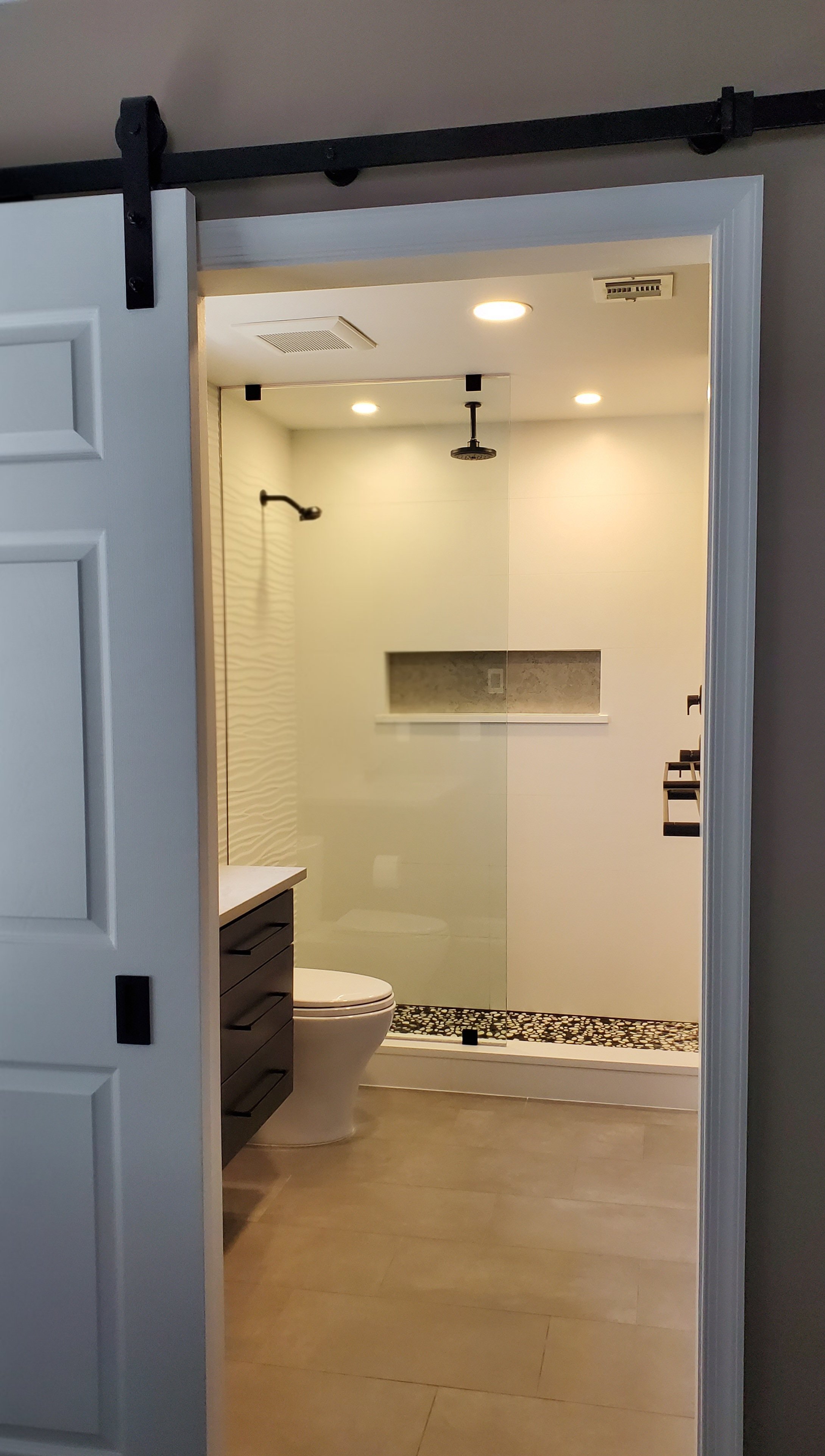 A black barn door slides open to a glass-walled walk-in shower with wave-textured white tile, a storage niche, and a pebble-tile floor.