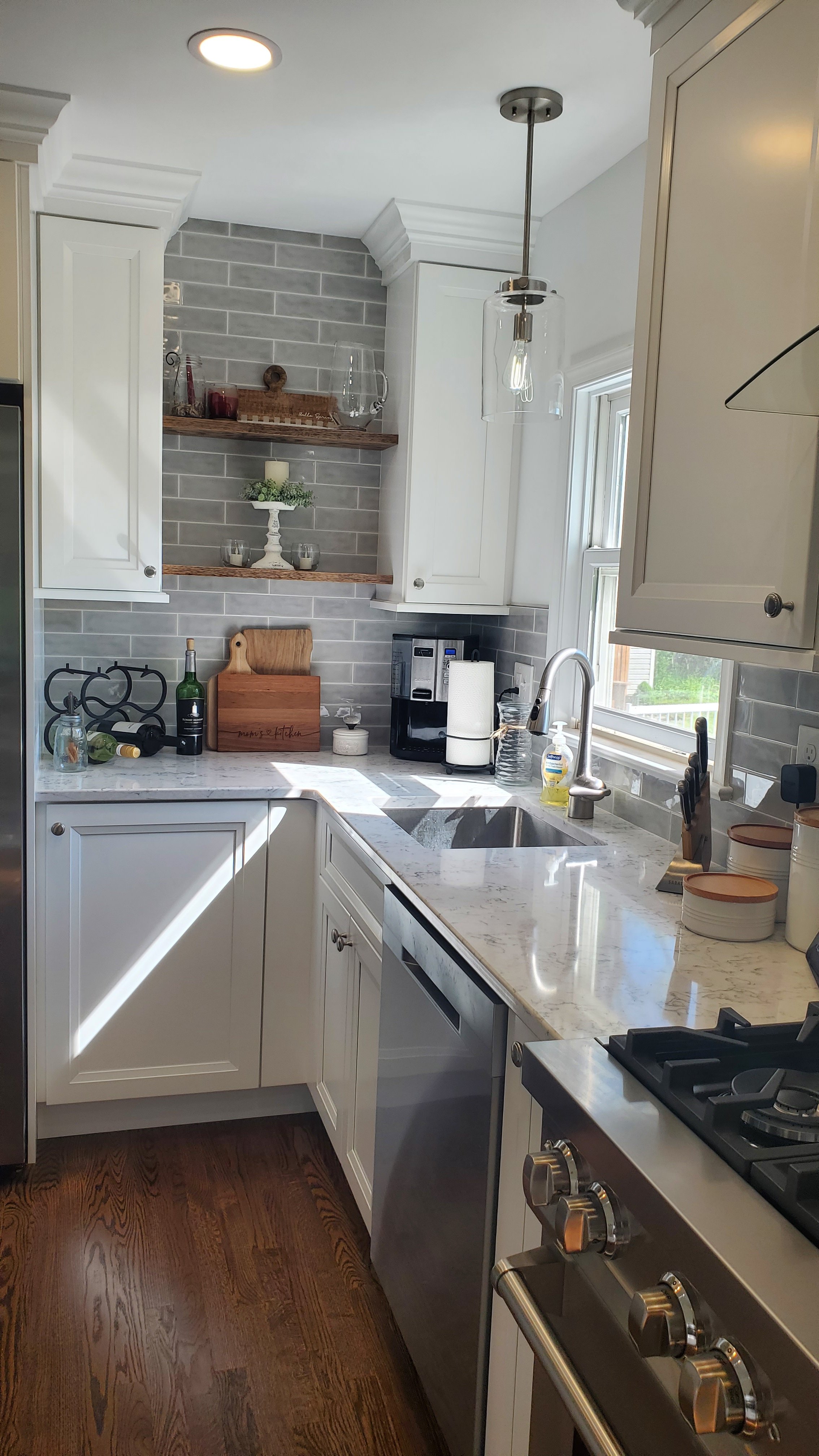 White shaker cabinets and marble-look quartz countertops sit on gray plank tile flooring, alongside a stainless steel gas cooktop and a wine rack near a window.