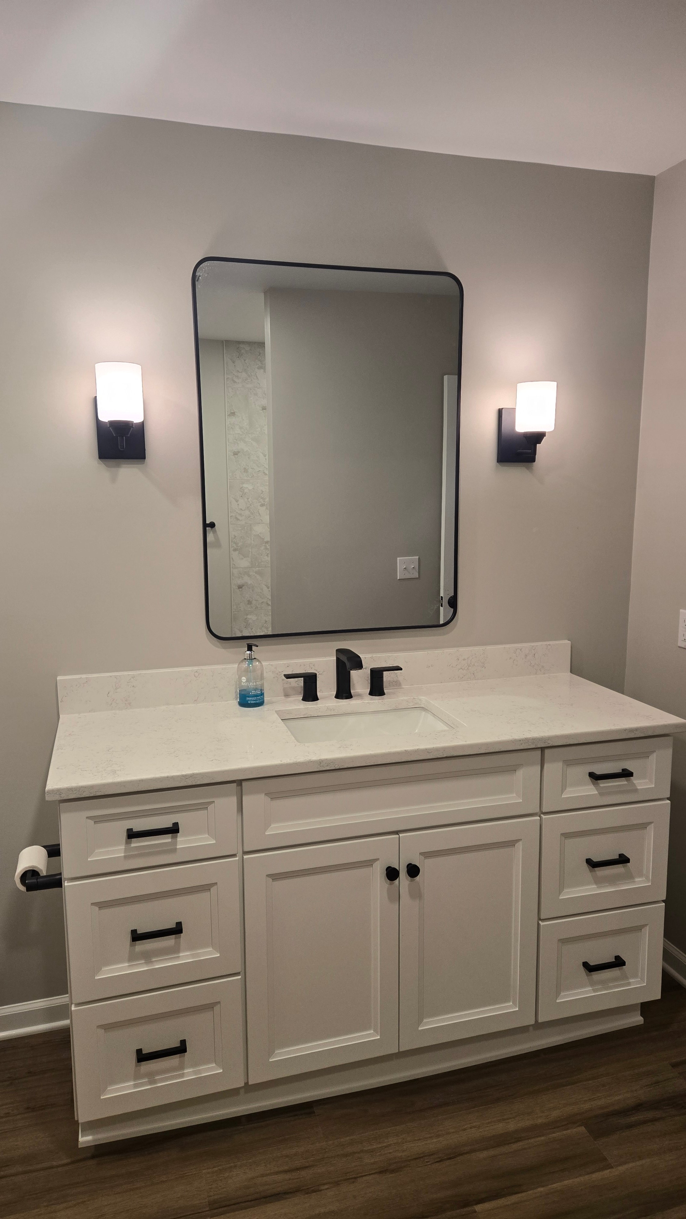 A white shaker-style vanity with black bar pulls and a matte black wall-mount faucet sits beneath a black-framed mirror flanked by black sconces.