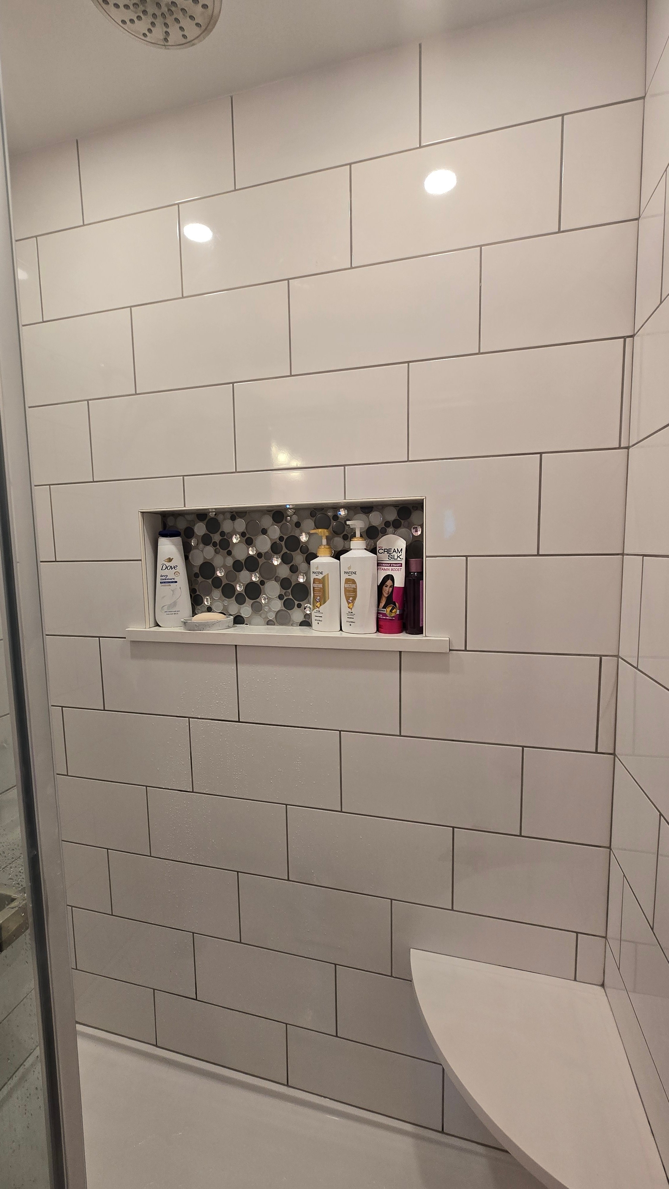 A recessed tiled shower niche lined with dark and light mosaic accent tile holds shampoo, conditioner, and body wash bottles, framed by staggered white subway tile.