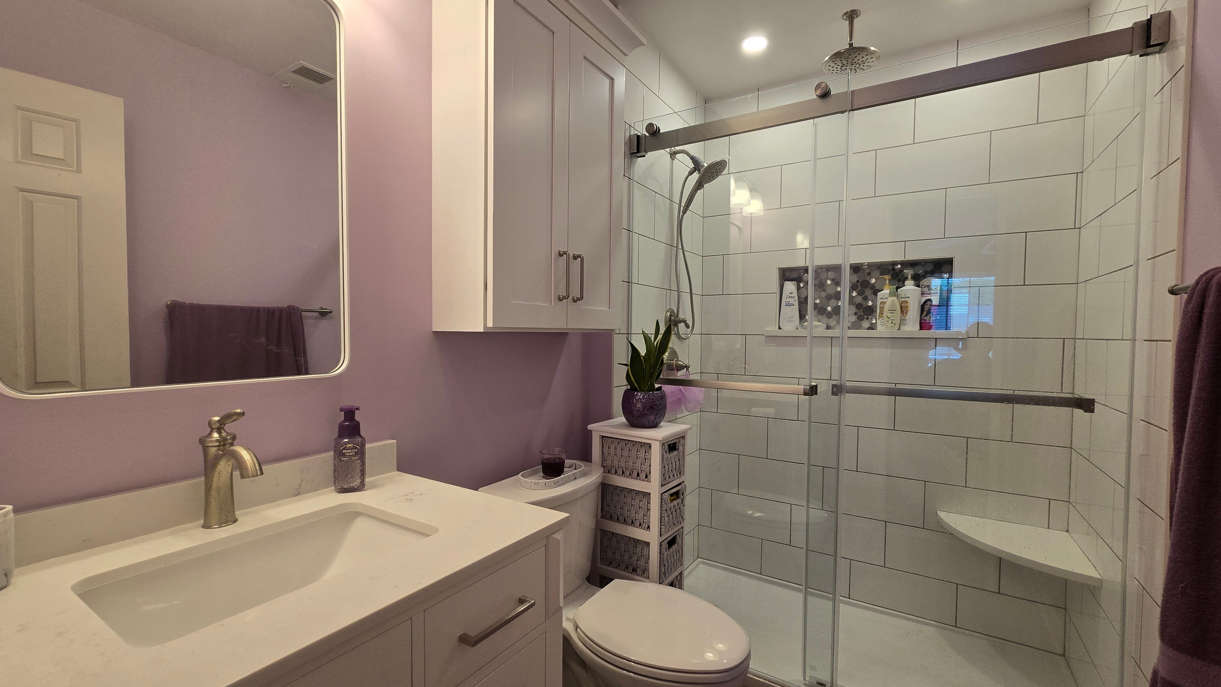 A white vanity with a marble-look quartz countertop, brushed nickel faucet, and round mirror stands against a lavender accent wall.