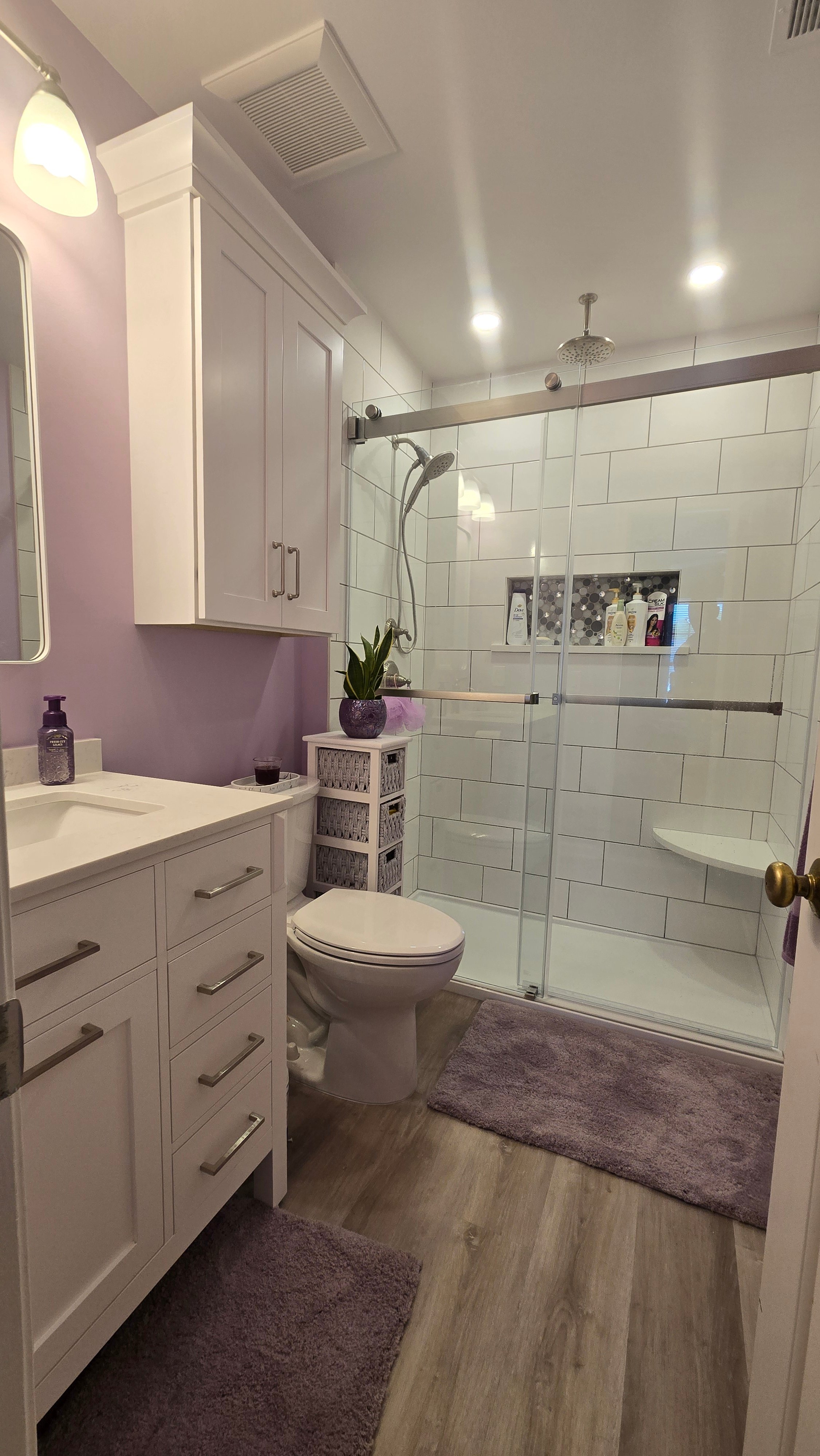 A glass-enclosed shower with white subway tile walls and a tiled niche pairs with a gray vanity, brushed nickel pulls, and a lavender countertop and accent wall.