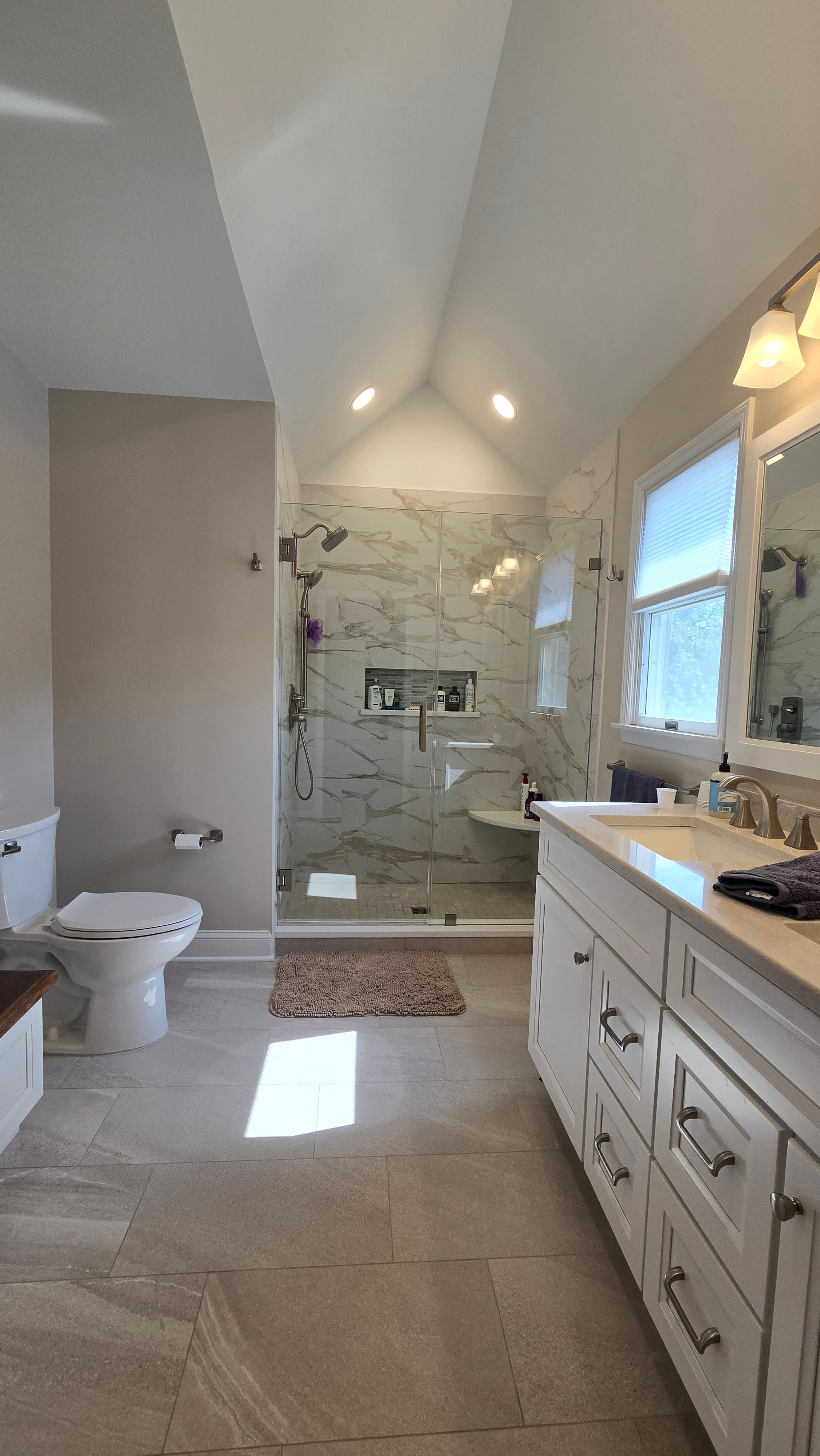 A walk-in shower has floor-to-ceiling marble-look porcelain tile, a frameless glass door, and a recessed tiled storage niche.