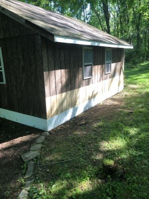 After photo of the same shed with new wood siding installed, showing a clean exterior with a partial coat of stain applied and a stone-lined drainage path along the foundation.