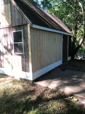 After photo of the repaired shed exterior, showing new siding, a finished roofline, and white trim, set against a wooded backdrop.