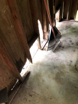 Before photo of the shed's interior showing exposed wall studs with rotted and damaged wood framing, with a rake and shovel leaning against the wall